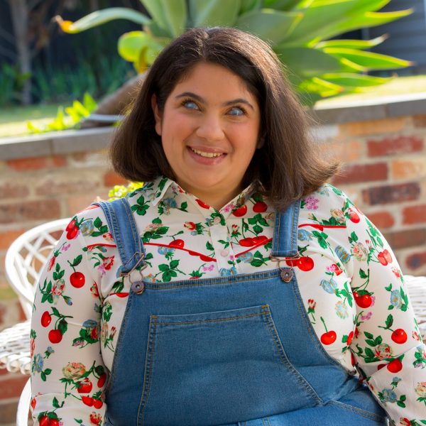 A smiling woman with shoulder-length brown hair, wearing denim overalls and a white collared shirt with a printed flower pattern. She is sitting in a garden, with a low brick wall, a white chair and a plant in the background.