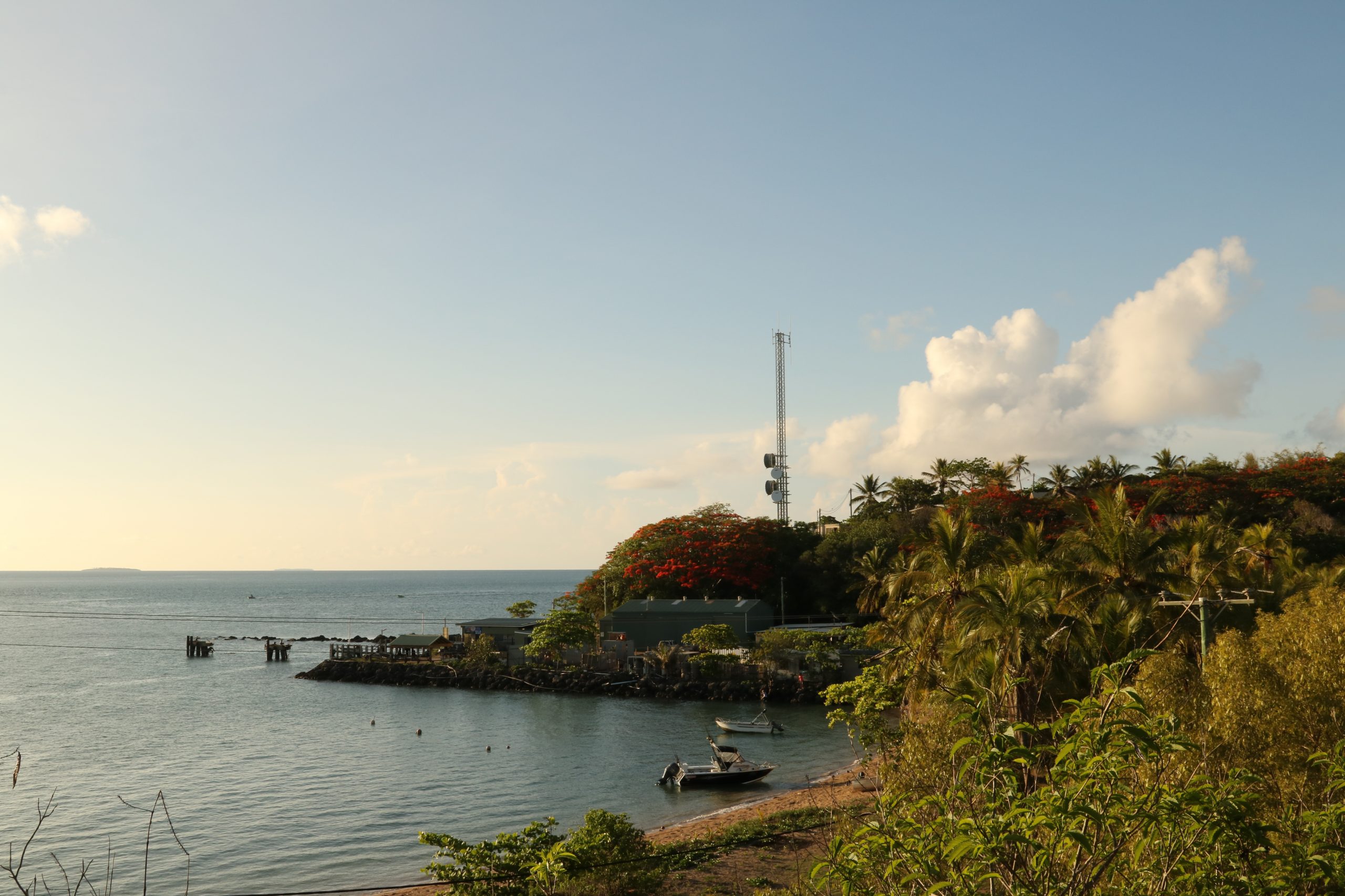 A small bay in the late afternoon, with palm trees in the foreground. On the far side of the bay is a metal shed under large poinciana tree and next to a telecoms tower. Next to the building is a wharf protruding out into the ocean. There are two small boats in the bay.