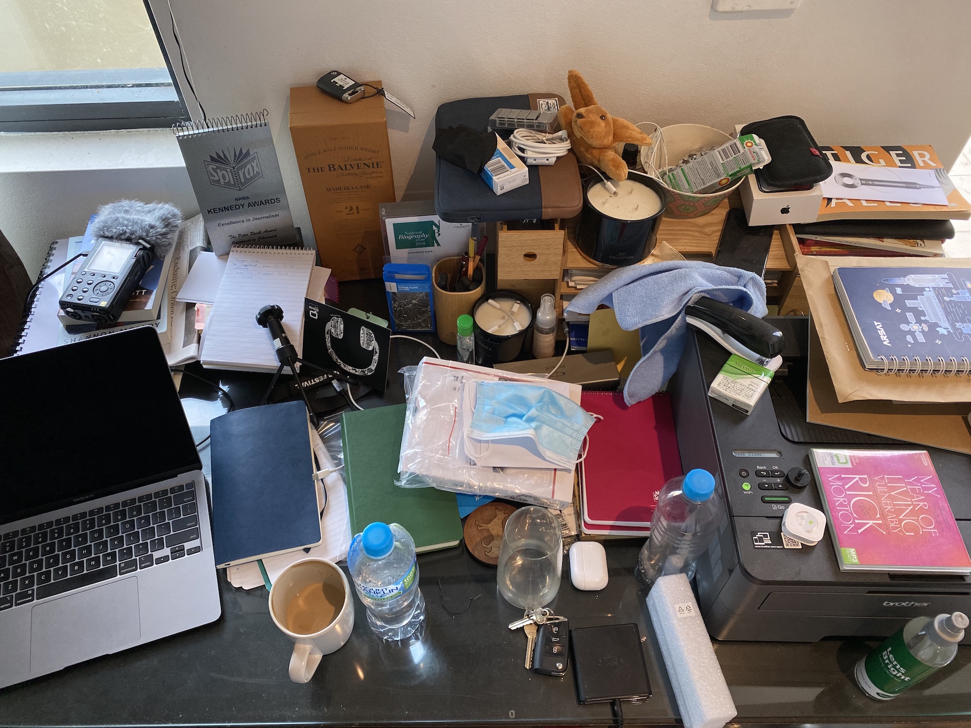 A photograph of a desk with a laptop in the bottom left corner, as well as cups, notebooks, cables and paraphernalia over the desk, and a printer in the bottom-right corner