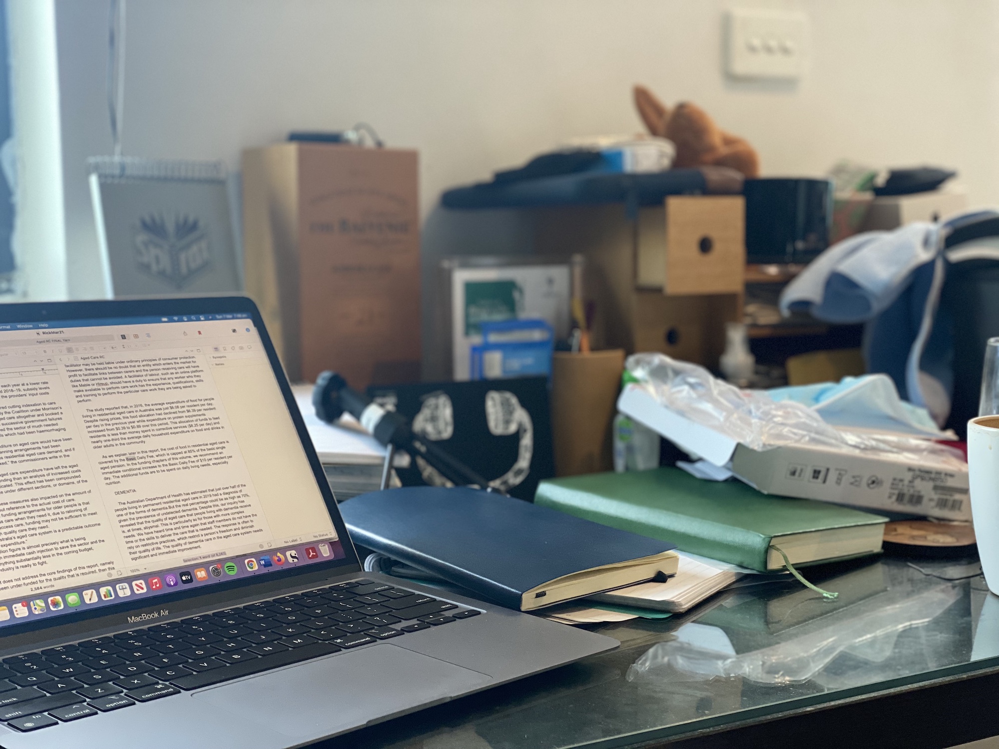 A desk with a laptop in the foreground, and the Scrivener writing program open onscreen. In the background the desk is covered in notebooks and other paraphernalia.