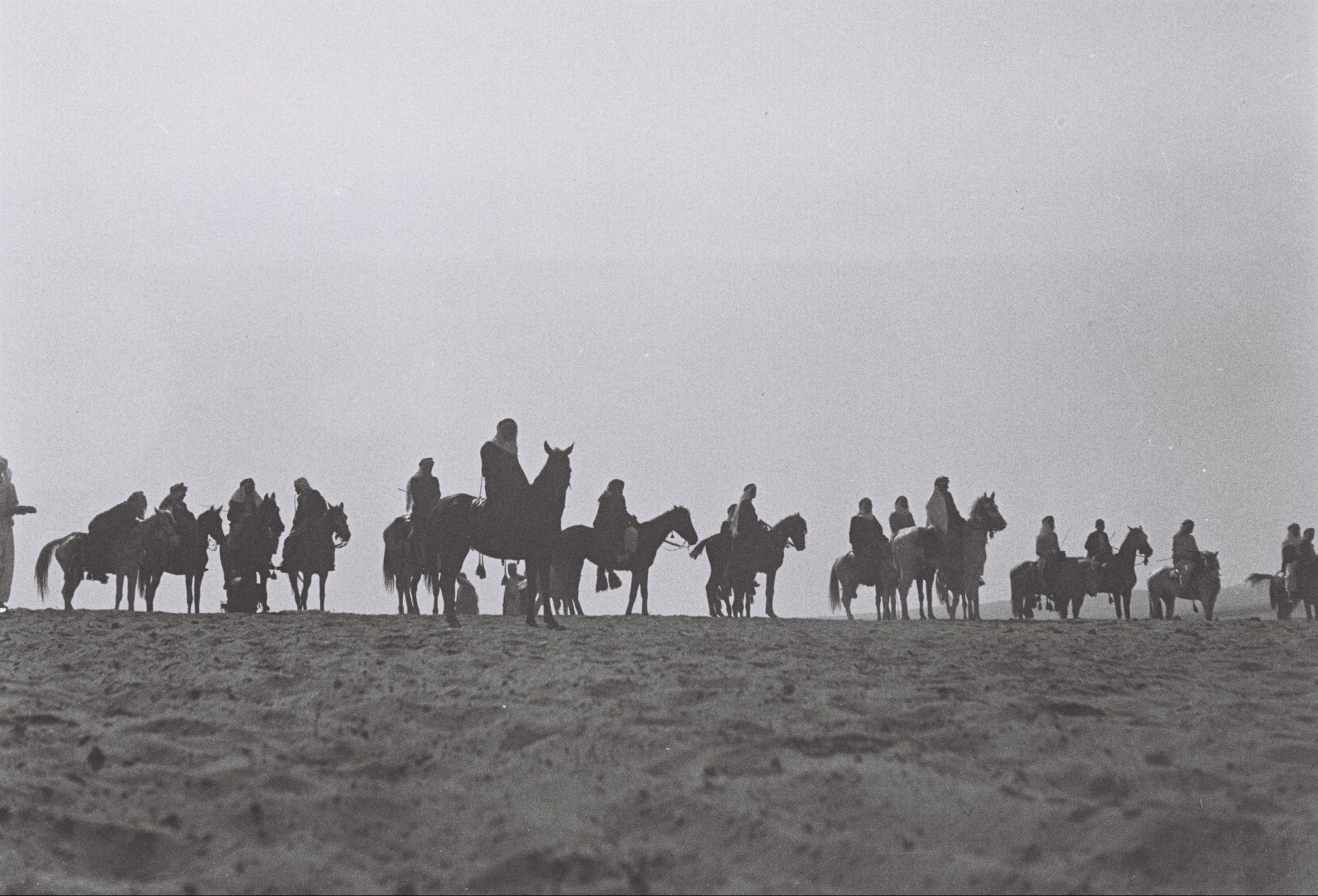 A black and white photo of Arab men in traditional clothing, on horses in the desert. The men are in the middle distance and mostly in silhouette.