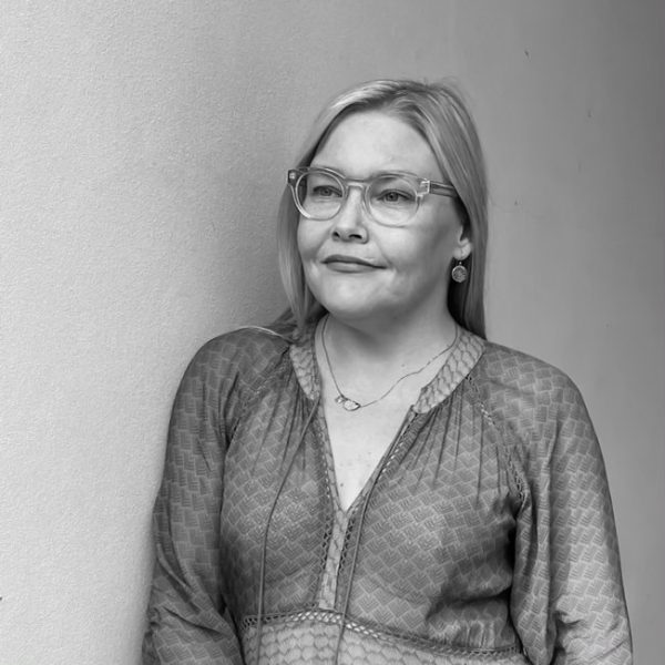 A black-and-white photo of a white woman with long blonde hair, leaning against a wall, looking to the right of the camera and wearing glasses, a thin necklace and a button-up top