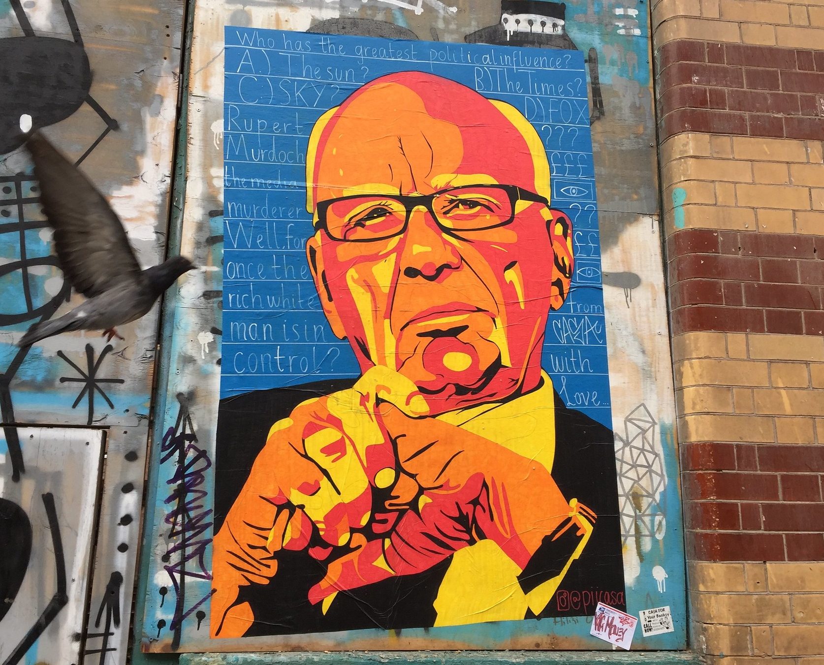 A street art stencil poster of Rupert Murdoch on a wall covered in other graffiti.