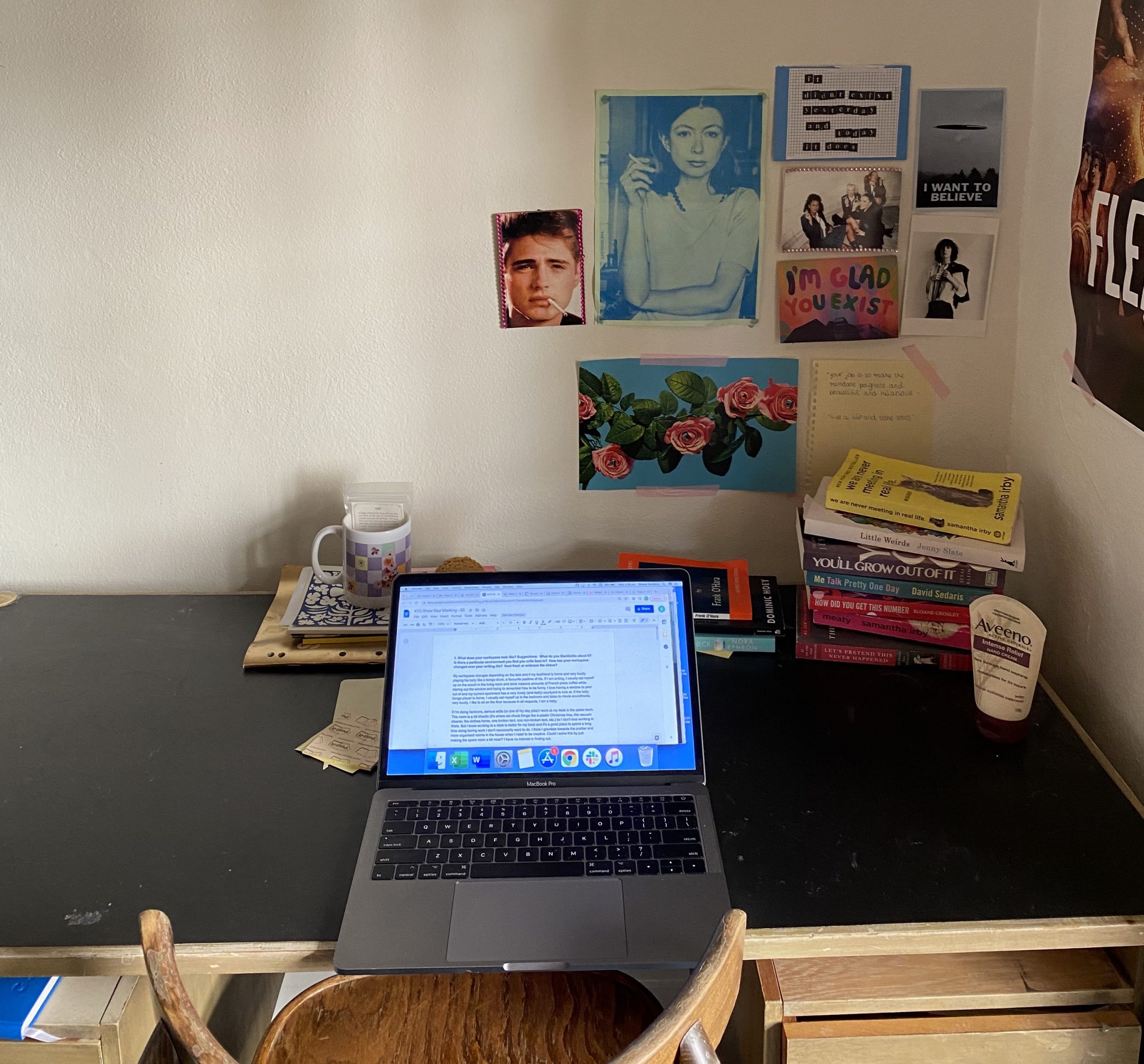 Sinead Stubbins' desk. An Apple laptop open to a Google Docs page on a timber desk with a black vinyl top. In the right back corner of the desk there is a pile of books and a bottle of moisturiser. Blue-tacked on the wall are various photographs and photos of flowers, women and pieces of text.