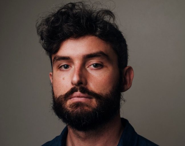 An image of Lech Blaine - a man with short dark brown hair, an olive complexion and a dark beard, wearing a blue shirt and looking at the camera with a neutral expression.