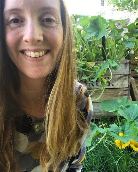 Lucinda Bain, a writer with long brown hair in a garden with green leaves and yellow flowers.