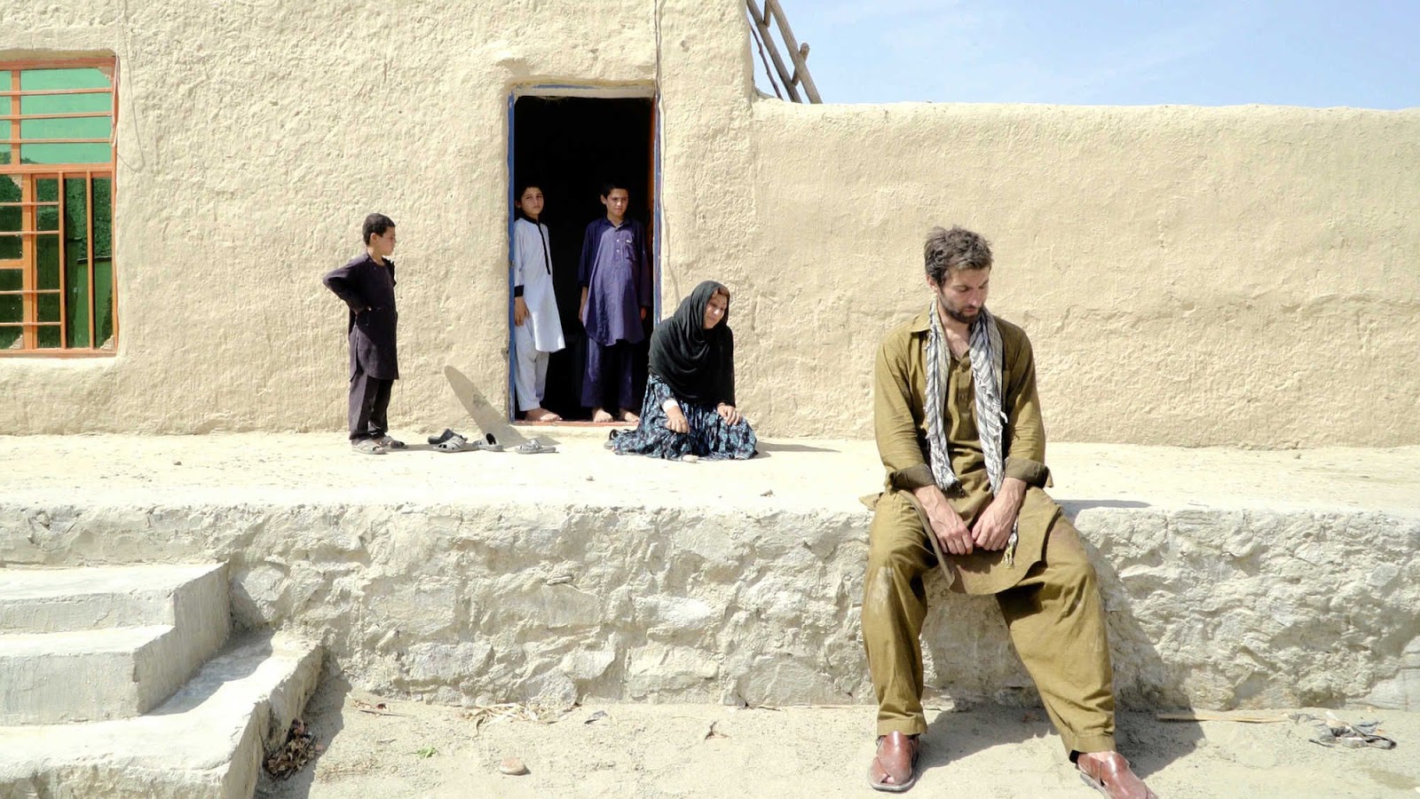 A still from the film Jirga: An Australian man wearing Afghan style clothes sitting on a ledge outside a white mudbrick style house. Behind him are a woman kneeling and three children looking on.