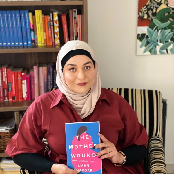 Amani Haydar, wearing a red shirt and cream-coloured headscarf, sits on a striped armchair holding her book, The Mother Wound. Behind her a bookcase and a painting of a potted plant are partially visible.