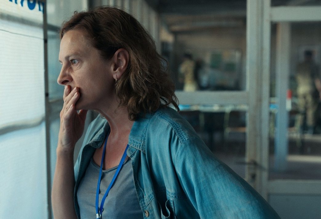 A still from the film 'Quo Vadis, Aida?'. Aida, a middle-aged woman with shoulder-length hair wearing a loose denim overshirt and a grey t-shirt, looks out a window with a concerned expression and her hand to her mouth.