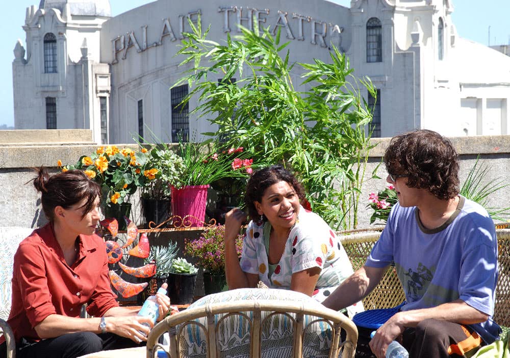 A still image from the Secret Life of Us. Claudia Karvan, Deborah Mailman and Samuel Johnson are sitting in the sun on bamboo outdoor chairs; a rack full of potted plants is behind them, and is the distance is St Kilda's Palais Theatre.
