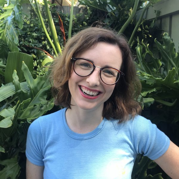 Brigid Mullane wearing a light blue t-shirt and glasses, smiling in front of a background of plants.