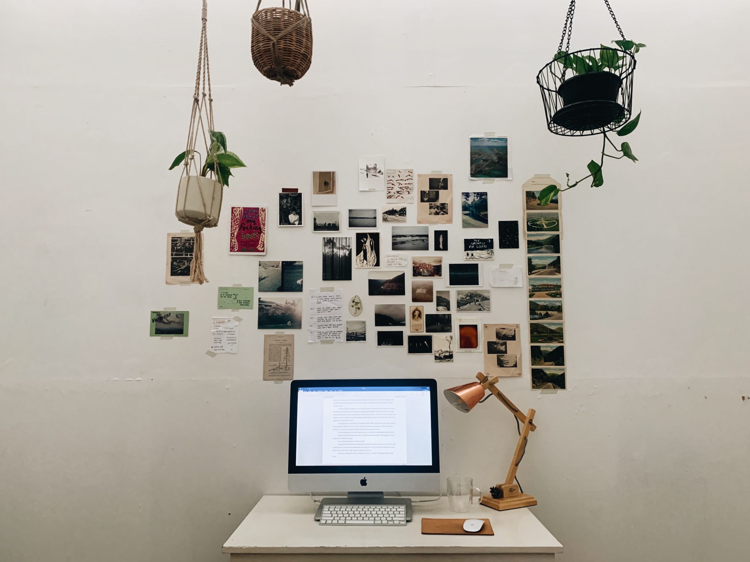 A silver iMac computer with an open Microsoft Word document on the screen, sitting on a white desk next to a timber and brass desk. Stuck to the white wall behind the desk are various photographs, postcards and other small items. Three small pot plants hang from the ceiling.