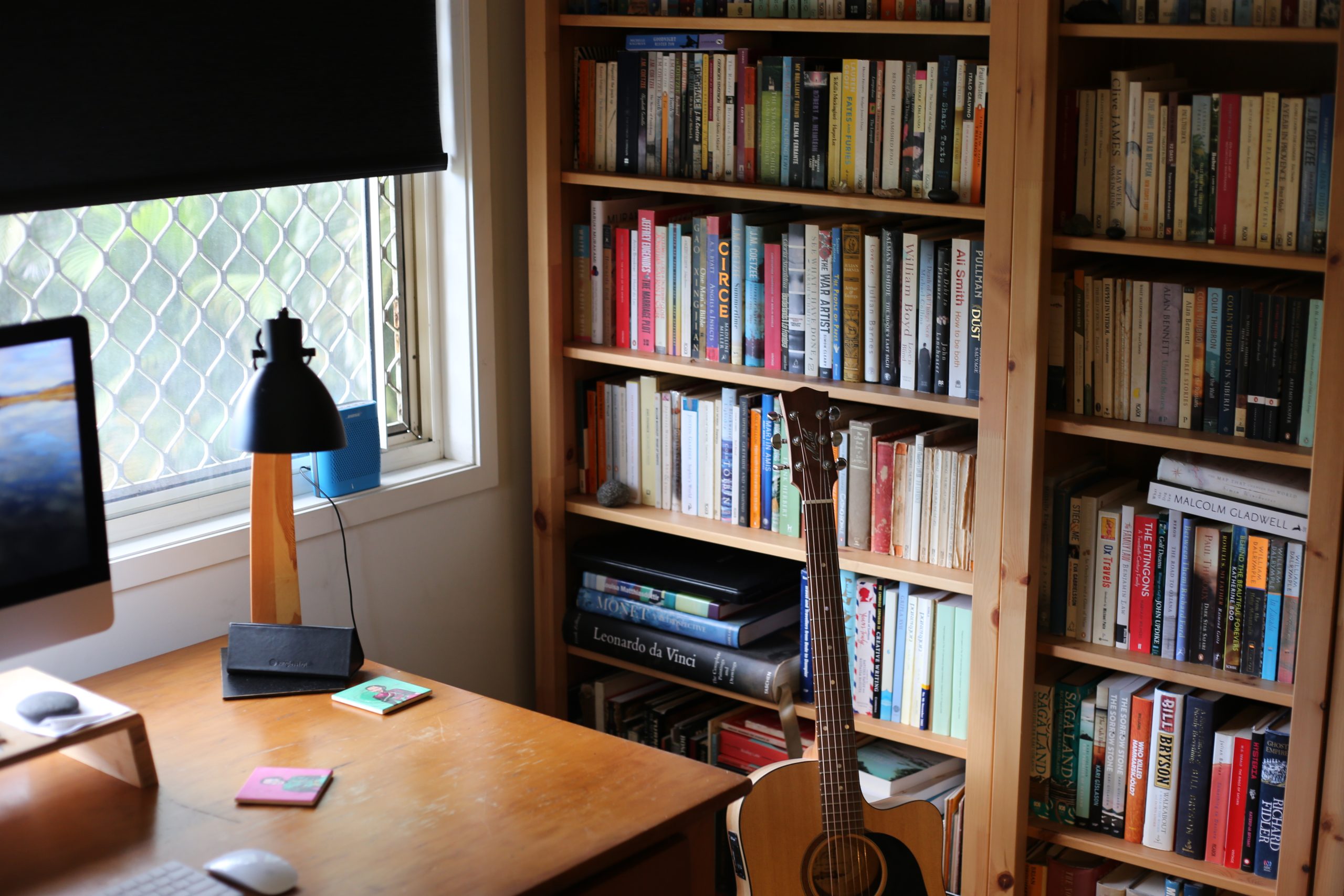 A light timber bookshelf filled with books next to a window and a timber desk with an iMac computer half out of frame. The desk is mostly empty except for a keyboard and mouse, two colourful drink coasters and a black and timber lamp. A guitar is propped up on the floor between the desk and the bookshelf. 