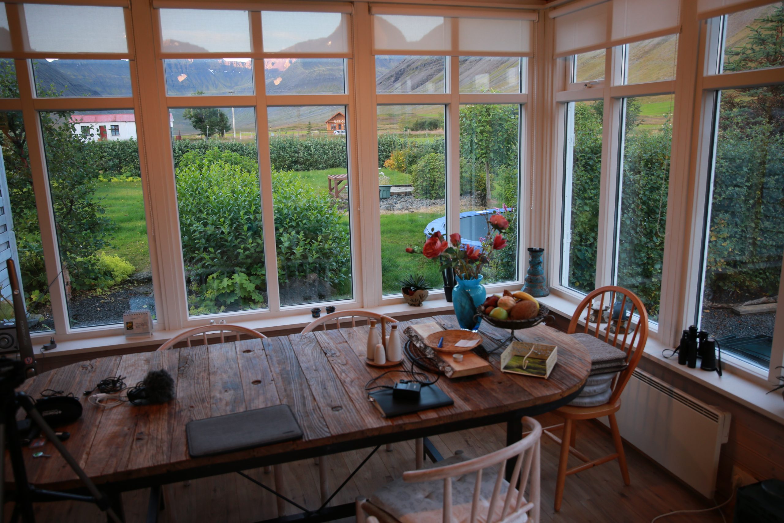 A large reclaimed timber dining table with four wooden chairs, in a corner of a room with full-length windows along two entire walls. On the table is a laptop, notebook, fruit bowl, vase of flowers, and various other small items. The view out the window is of a lush green orchard with farm sheds and large mountains in the distance.
