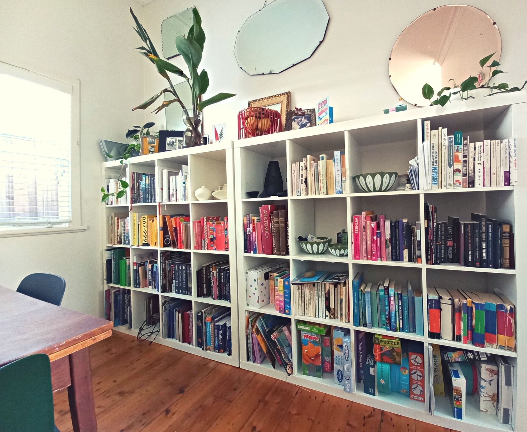 A white bookshelves extending horizontally along a wall and halfway up it. The shelves are filled with books organised according to colour, as well as some board games and pottery. Above the bookshelf is 3 large wall-mounted mirrors, several indoor plans, photographs and other objects.