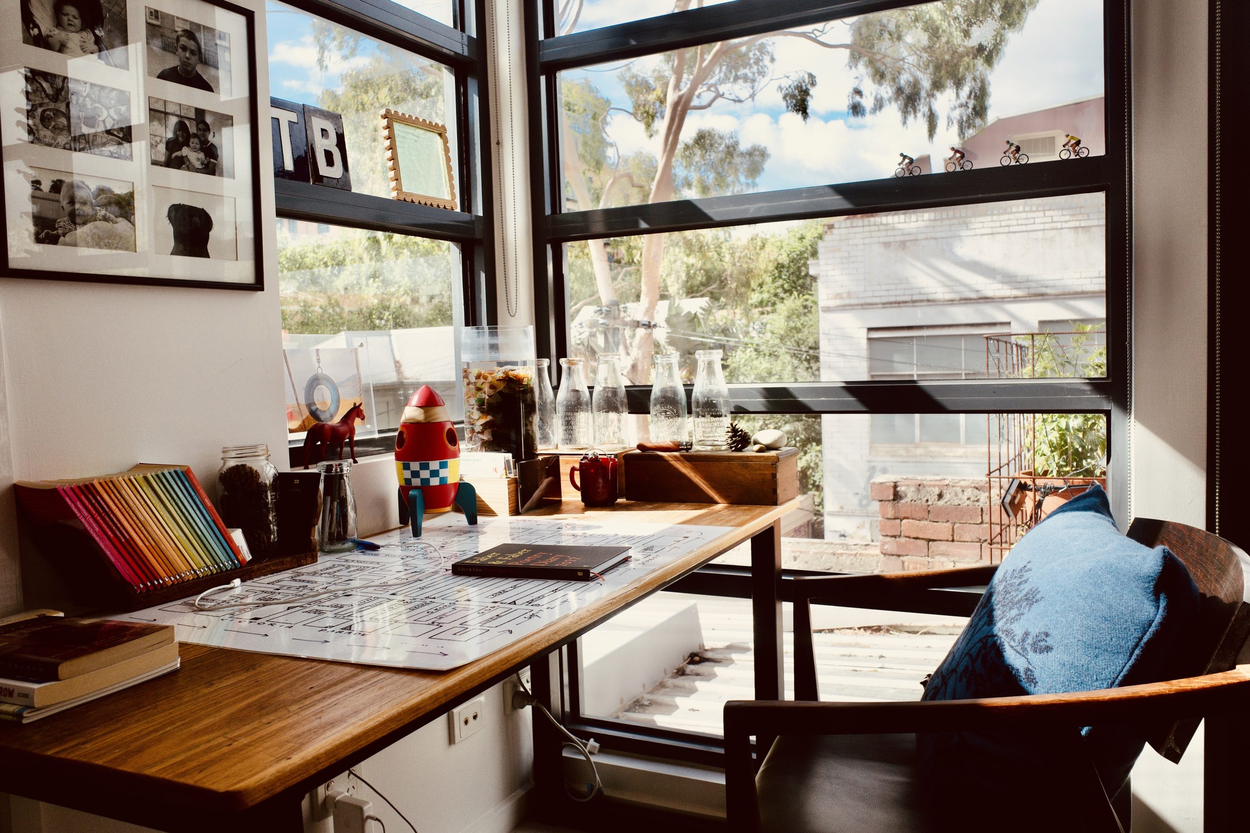 A large timber desk next to a large bright corner window. On the desk is a large white placemat with a vintage street map of Fitzroy printed on it, as well as an assortment of jars and other trinkets, a toy horse and rocket, a series of books with their spines out in the colours of a rainbow. Outside the window a large eucalyptus tree is visible.