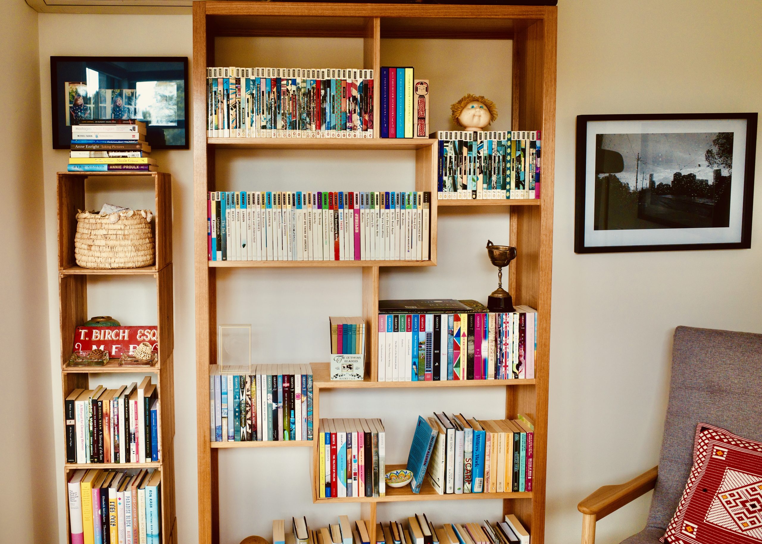 A full length timber bookshelf against a tan coloured wall. The shelves are cut into alternately-spaced squares, and are full of neatly organised journals and other books. A second, smaller set of shelves beside the main bookshelf has a woven basket and various other items on the shelves.