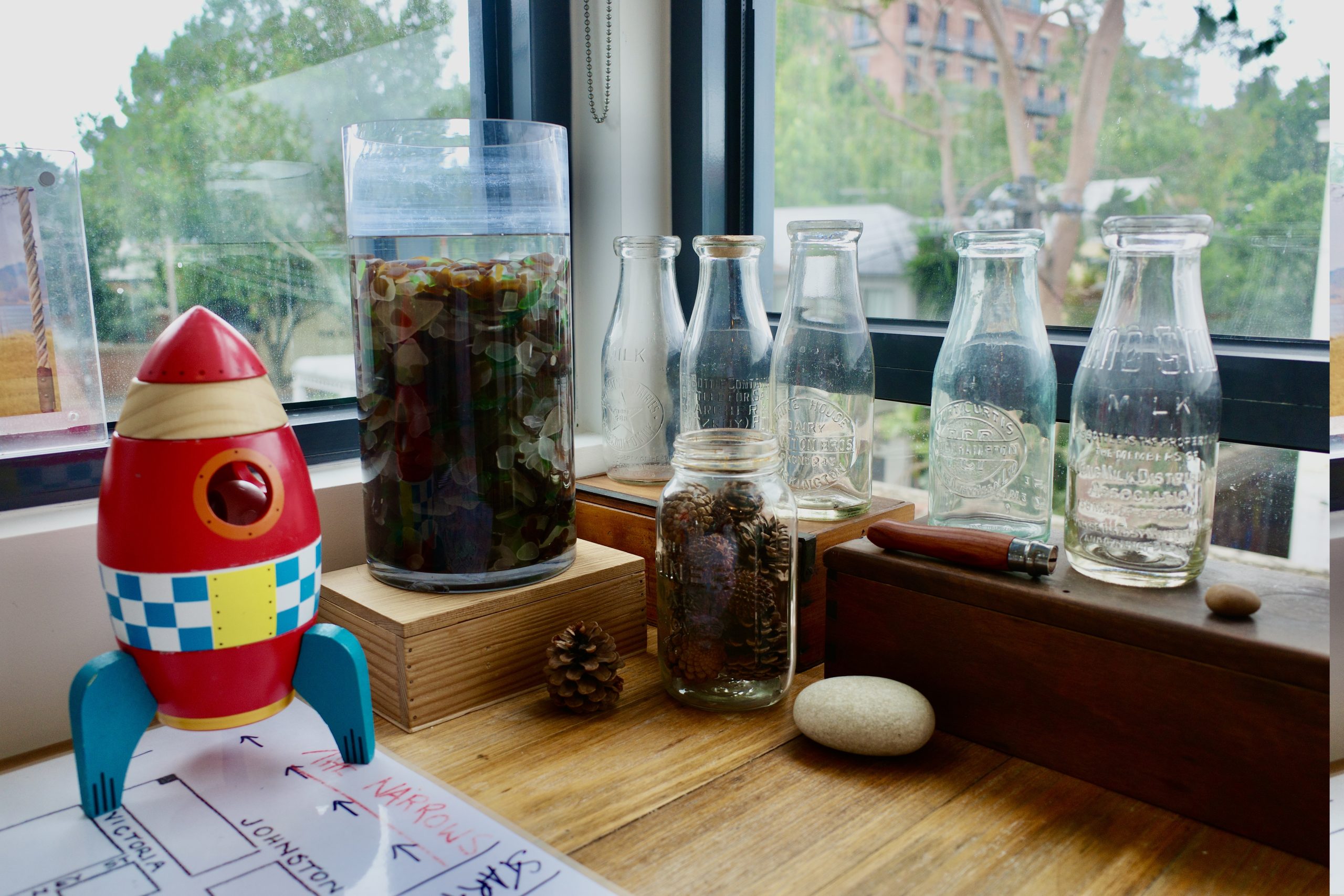 A selection of empty vintage milk bottles, a jar full of pine cones, a red and blue toy rocket, a jar full of colourful pebbles and a white stone, sitting on the corner of a timber desk.