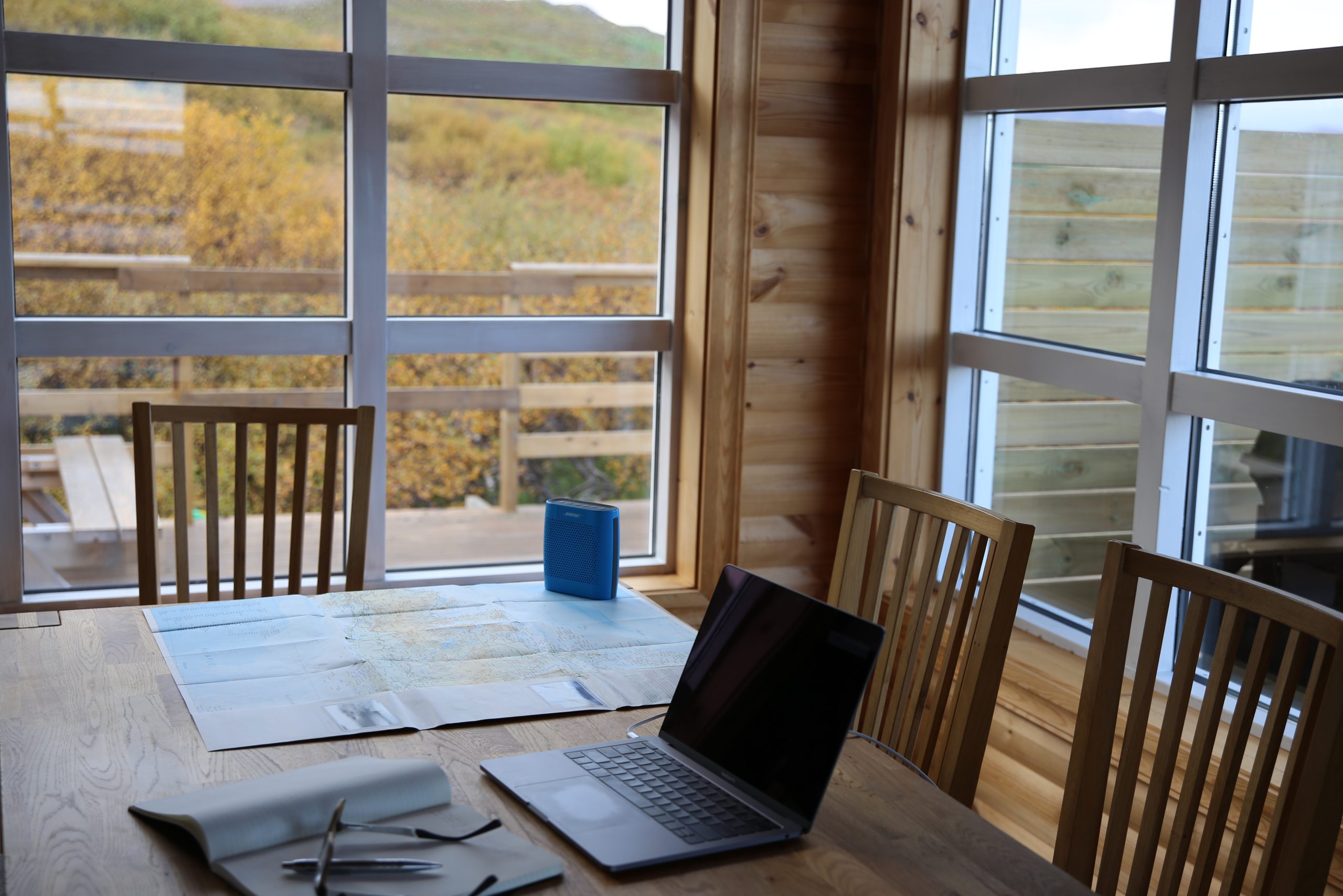 A light timber dining table with a laptop, notebook, map and small blue Bluetooth speaker. Around the table are three timber chairs in the corner of a room with similarly light timber walls. On each wall is a large white-framed window, and outside the windows is a grassy hillside paddock. 