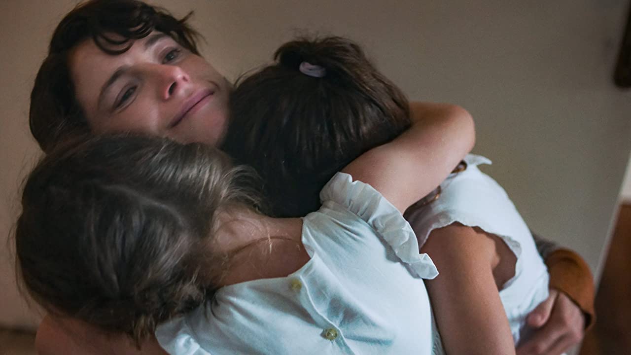 A shot from The Lost Daughter. Jessie Buckley, a young woman with short dark hair, hugs two you children in light blue dresses. She is smiling but with a slightly blank expression.