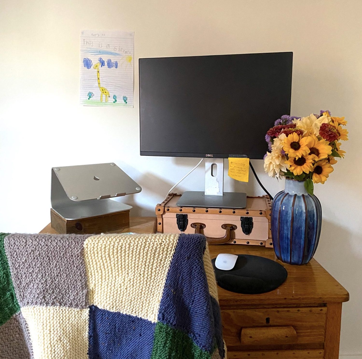 A small timber desk against a light-coloured wall. On the desk a silver laptop stand sits on a small wooden box and a large Dell external monitor with an orange sticky note attached to the bottom sits on a light brown suitcase-style box. Also on the desk are a mouse on a black mouse pad and a blue ribbed vase full of bright-coloured flowers. Over the desk chair is drape a knitted blue, green, off-white and grey blanket.