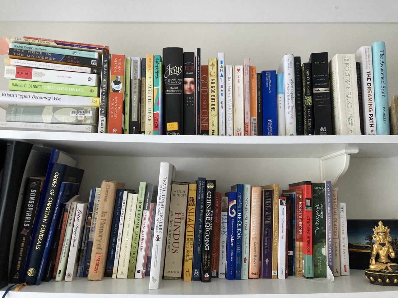 Two shelves of a white melamine bookshelf filled with books, including several titles on different religions. In the bottom right corner is a small golden statue of a seated Asian god.