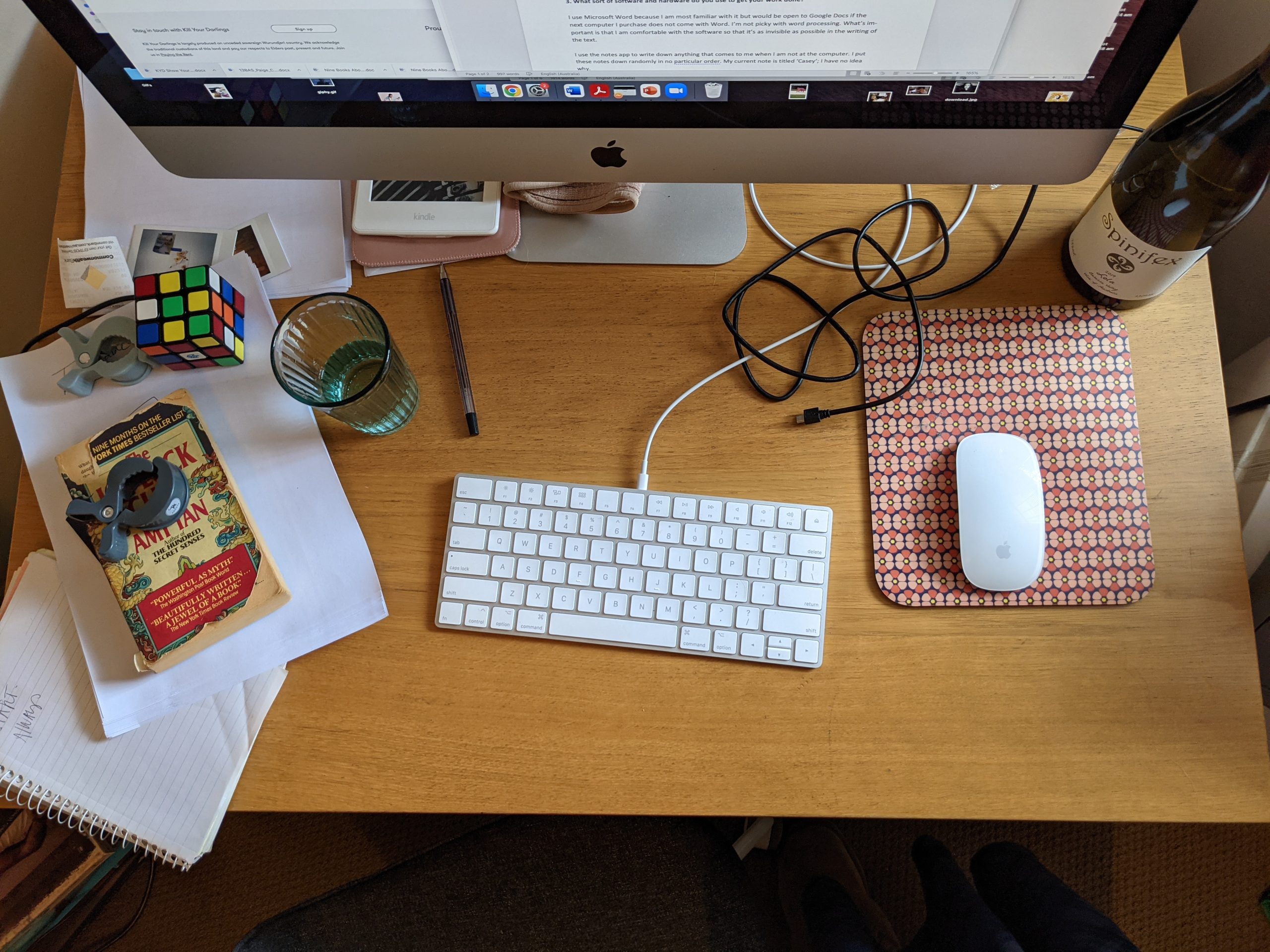 a top-down view of a timber desk with a white Apple keyboard and mouse, strewn with papers, books, cables and other items such as cups and a Rubik's cube.