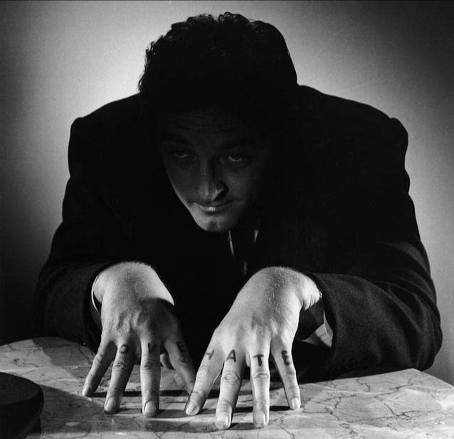 A black and white still image from The Night of the Hunter. A man in a black suit, bathed in shadow, sits at a table with his hands perched oddly. On each knuckle are tattooed the words HATE and LOVE.