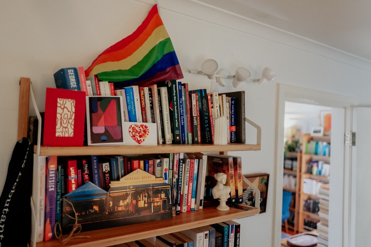 Two timber bookshelves mounted to a wall, filled with books, greeting cards, a rainbow flag and other assorted items.