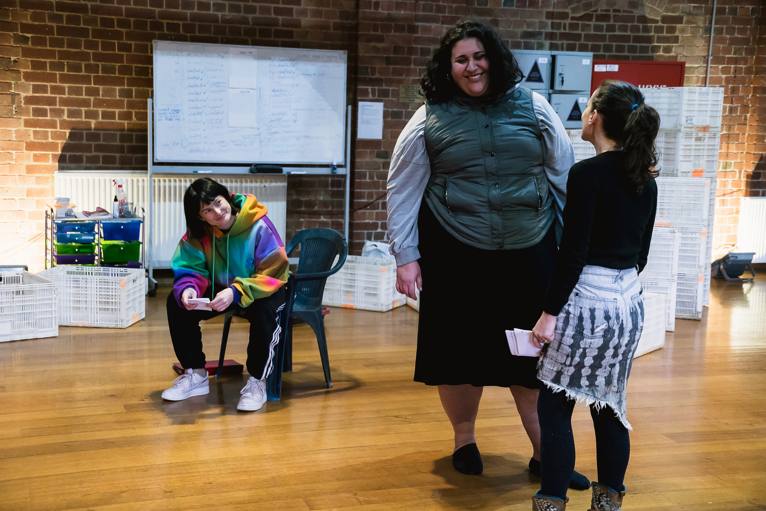 three people in a rehearsal room with timber floors and brick walls, surrounded by a whiteboard and other containers. Hannah Monson is sitting in a plastic chair, wearing a brightly coloured hoodie and smiling at Chanella Macri and Lucia Mastrantone, who are standing facing one another. Chanella is facing the camera and is laughing.