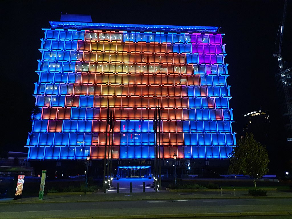 The side of an office building lit up at night, its windows forming a grid pattern on which the shape of an elephant is formed in orange against a blue background, with a pink love-heart shape in the top right corner