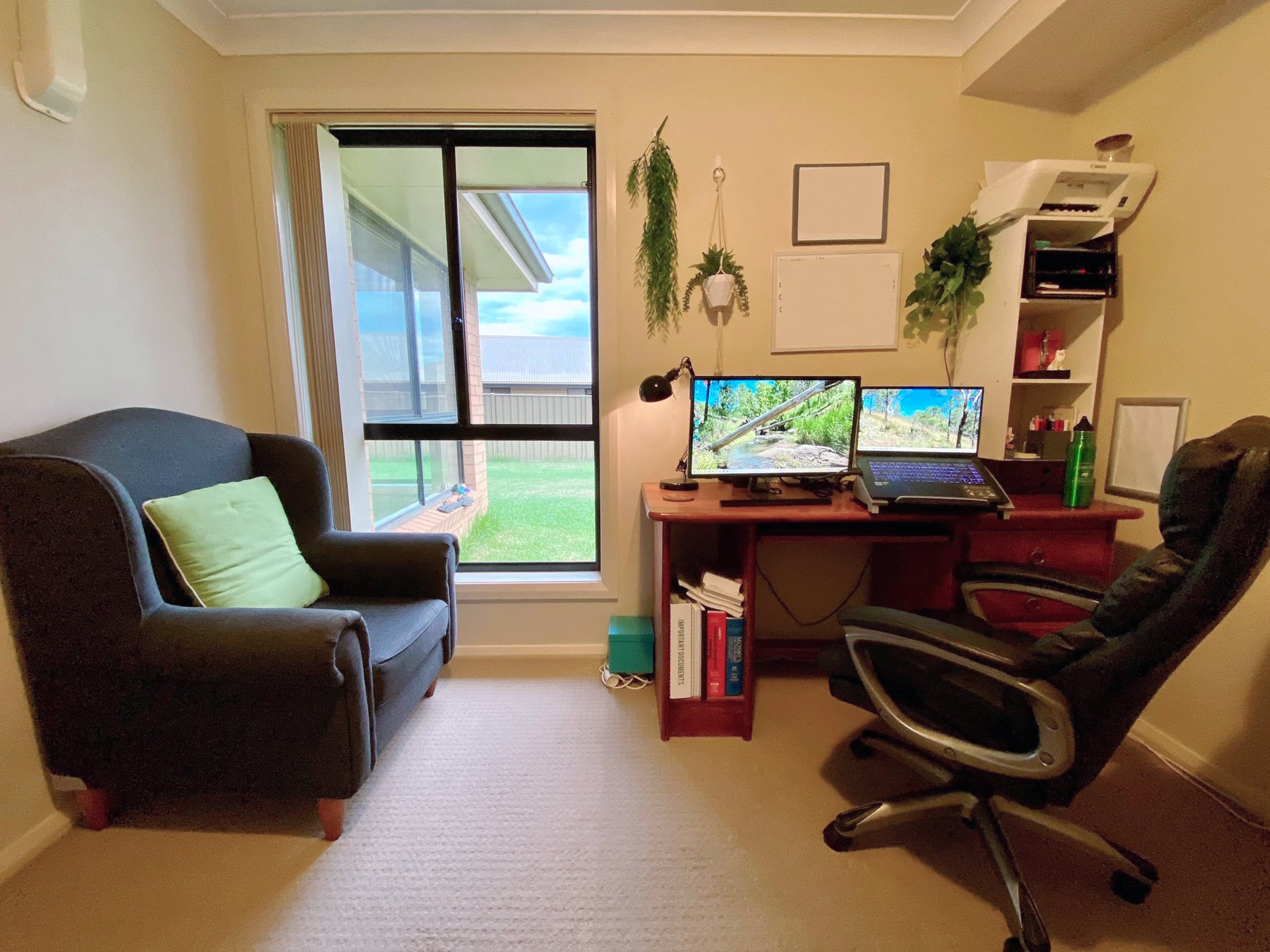 A large room with cream walls, cream carpet and a window on one wall opening out onto a lawn. A black armchair sits beside the window. Against the wall next to the window is a brown timber desk with a laptop on a stand, a monitor, and a whiteboard and assorted potplants mounted on the wall above. In front of the desk is a large black office chair.