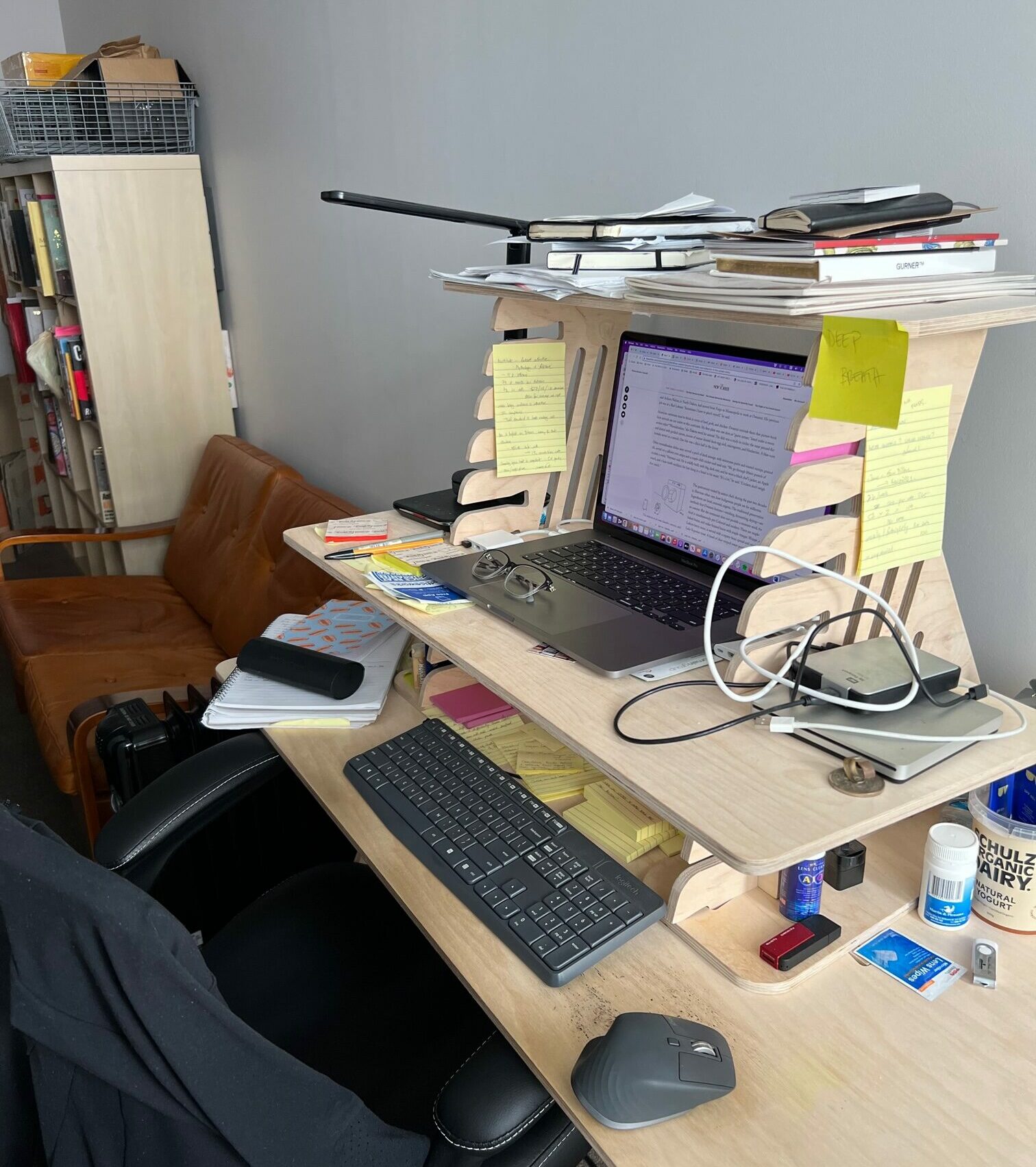 Jess Ho's workspace: A plywood three-tiered desk with an Apple laptop sitting on it with a word processor onscreen. On the top tier of the desk above the laptop are various papers and notebooks. On the lower tier is a keyboard and mouse and various cables, computer accessories, stationery and other items.
