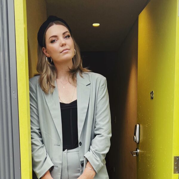 Amy Thunig standing and leaning against a yellow door frame. She is wearing a light grey suit with a black top underneath