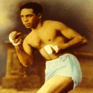 A vintage photograph of An Aboriginal man with short dark hair, wearing white shorts and white strapping around his fists, which are raised in a boxing stance.