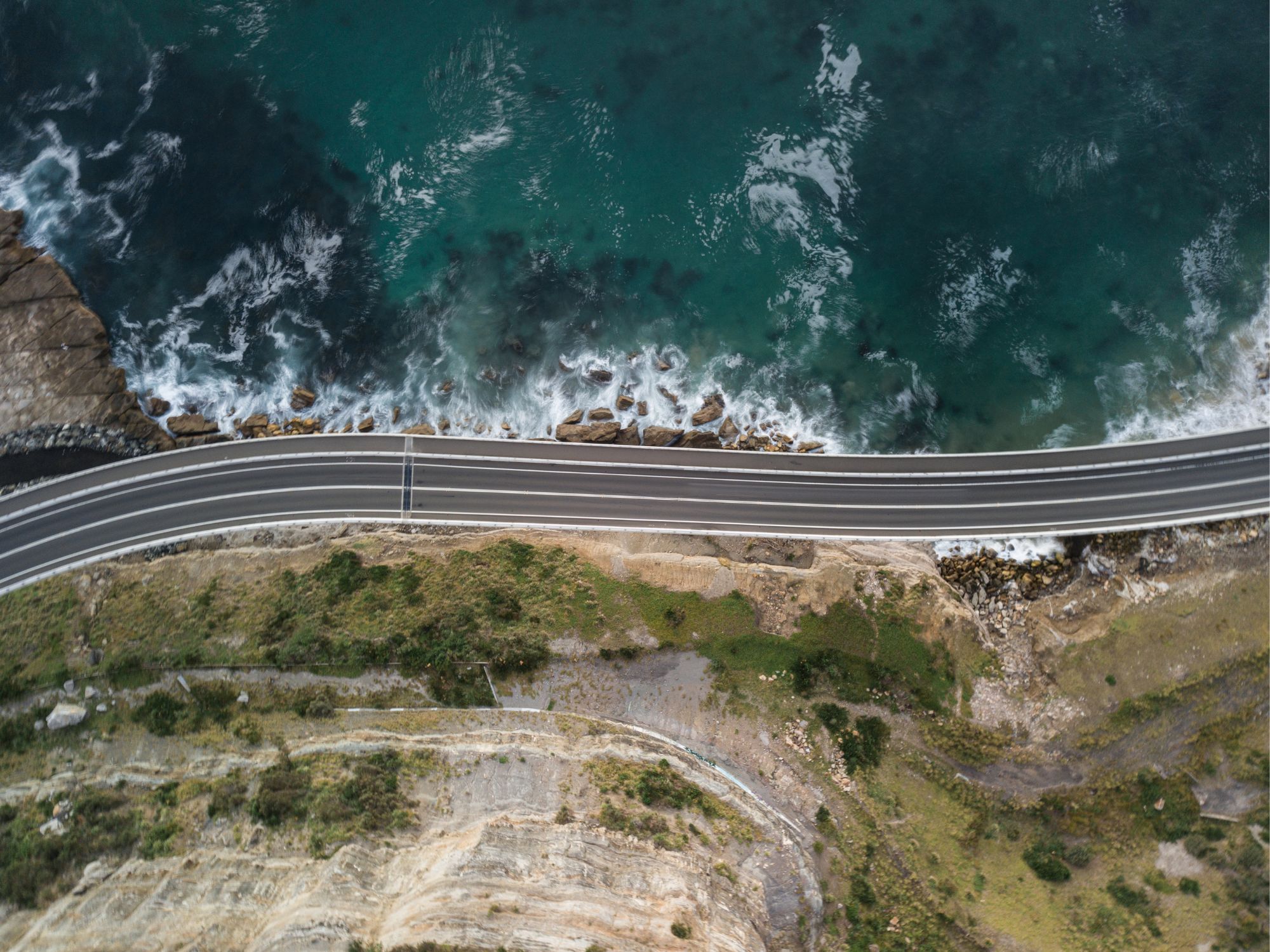 An overhead photograph of a road winding along a coastline. One one side of the road are grassy sandstone hills and on the other is the ocean.
